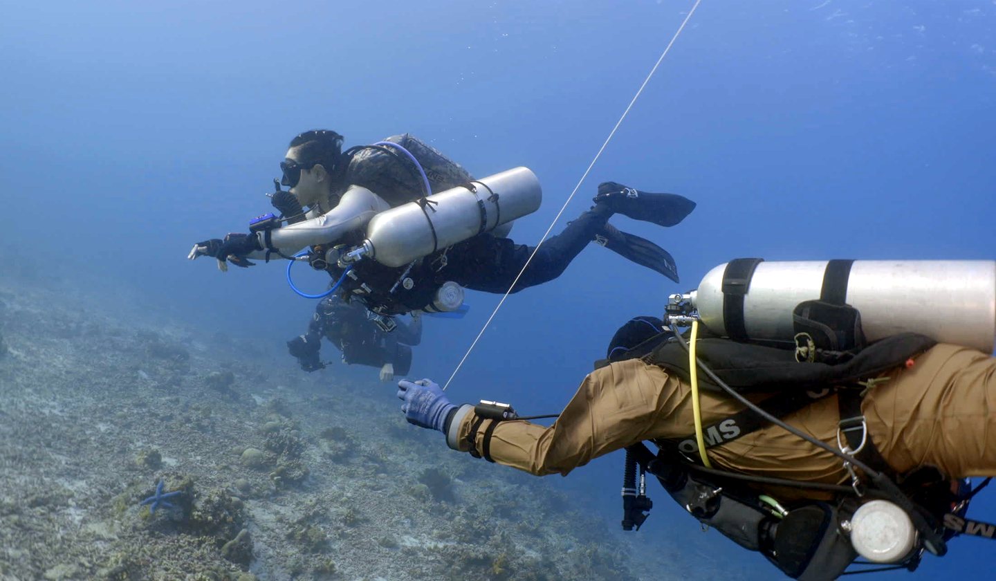 Two technical divers with twin cylinders and stage tanks swimming over the reef
